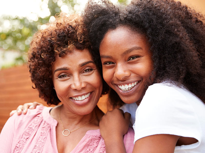 Middle aged black mum and teenage daughter embracing and smiling to camera